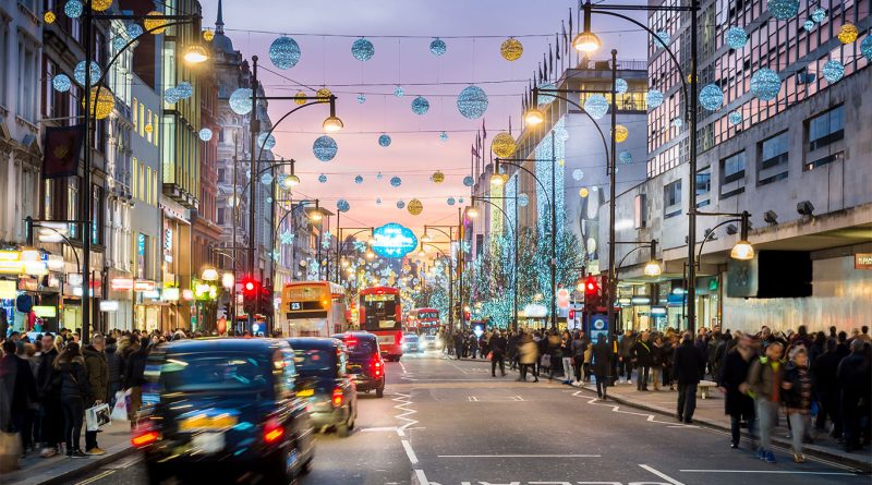 Image of Oxford Street in London displaying all the different types of shops, red buses, people walking on the sidewalk and lights hanging in the street