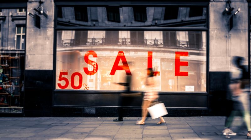 A busy UK high street with shoppers and retail stores displaying sale signs, reflecting the low inflation rates driven by falling clothing and footwear prices.