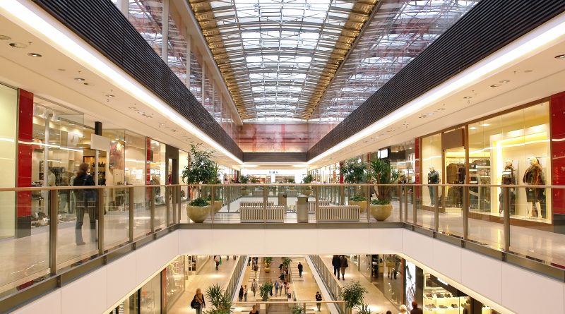 Young shoppers walking through a modern shopping mall while using smartphones to compare products and prices.