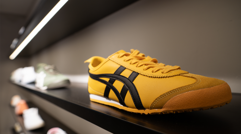 Close-up of a yellow and black Onitsuka Tiger sneaker displayed on a retail shelf, with other footwear styles softly blurred in the background under modern store lighting.
