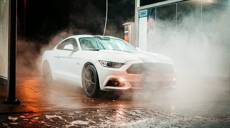 A white sports car drives through an automatic car wash, surrounded by mist and spraying water, with wet pavement reflecting warm orange lights.