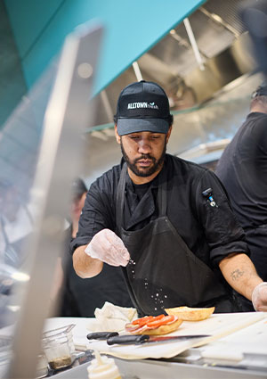 a kitchen employee wearing a black uniform and cap preparing food behind a counter