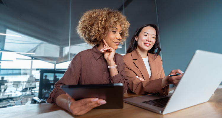 Two professional women are collaborating at an office desk, reviewing data on a laptop and tablet