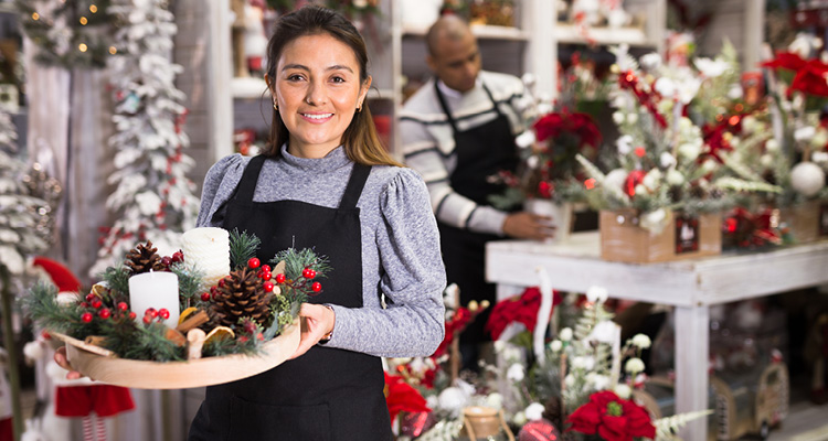 a smiling worker in a black apron holding a round Christmas centerpiece with white candles, pinecones, and red berries inside a festive retail or floral shop